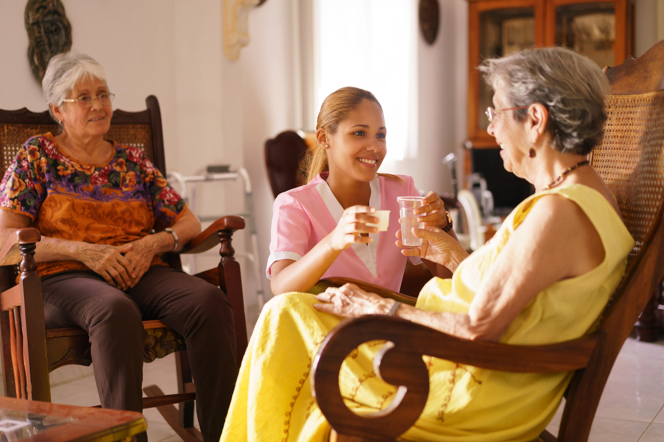 respite-care-in-Pipersville-PA-2 A smiling caregiver offers medication and water to a senior woman while another older adult looks on in a warm home setting.